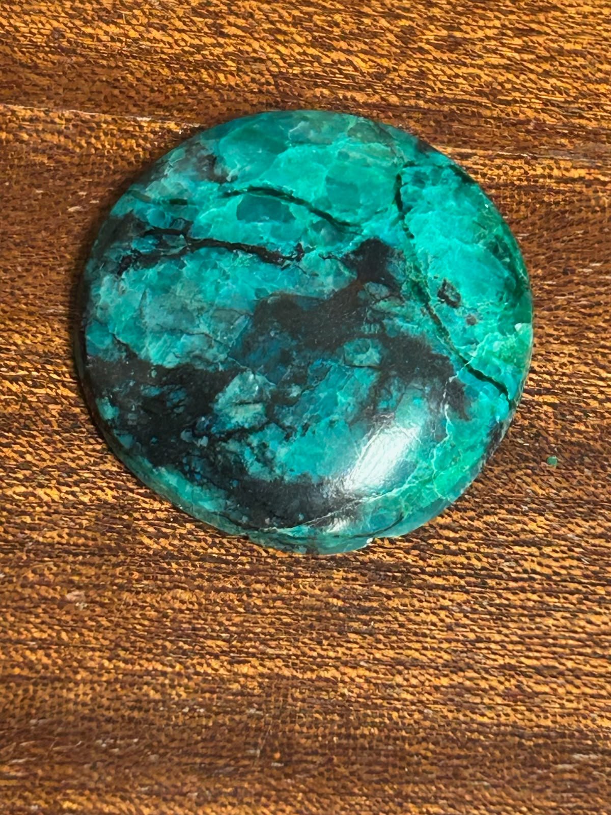 A round cabochon in blue green, veined and patched with black, on a wooden bench top.