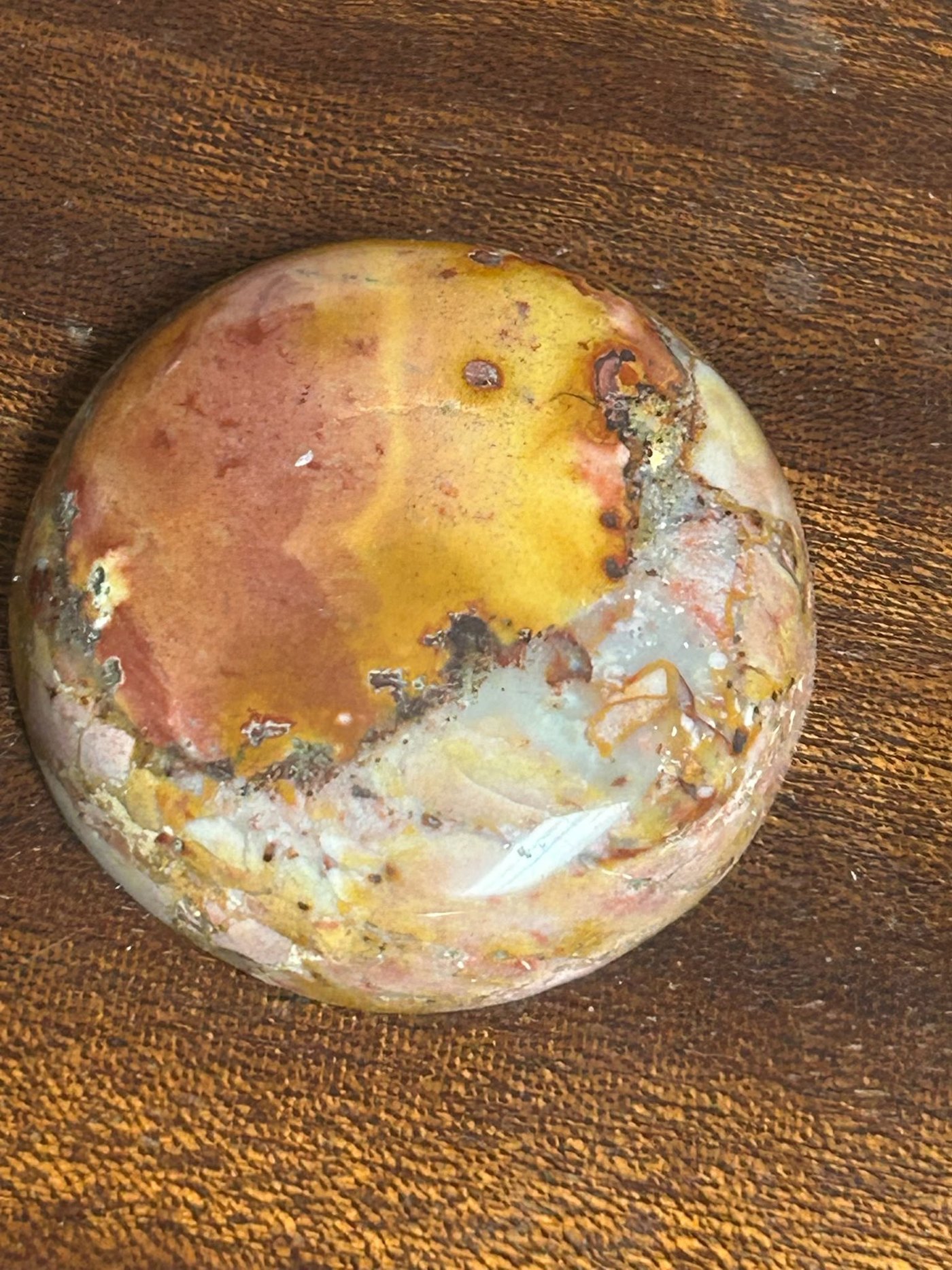 A round jasper cabochon in mustard, rust and cream, on a wooden bench top.