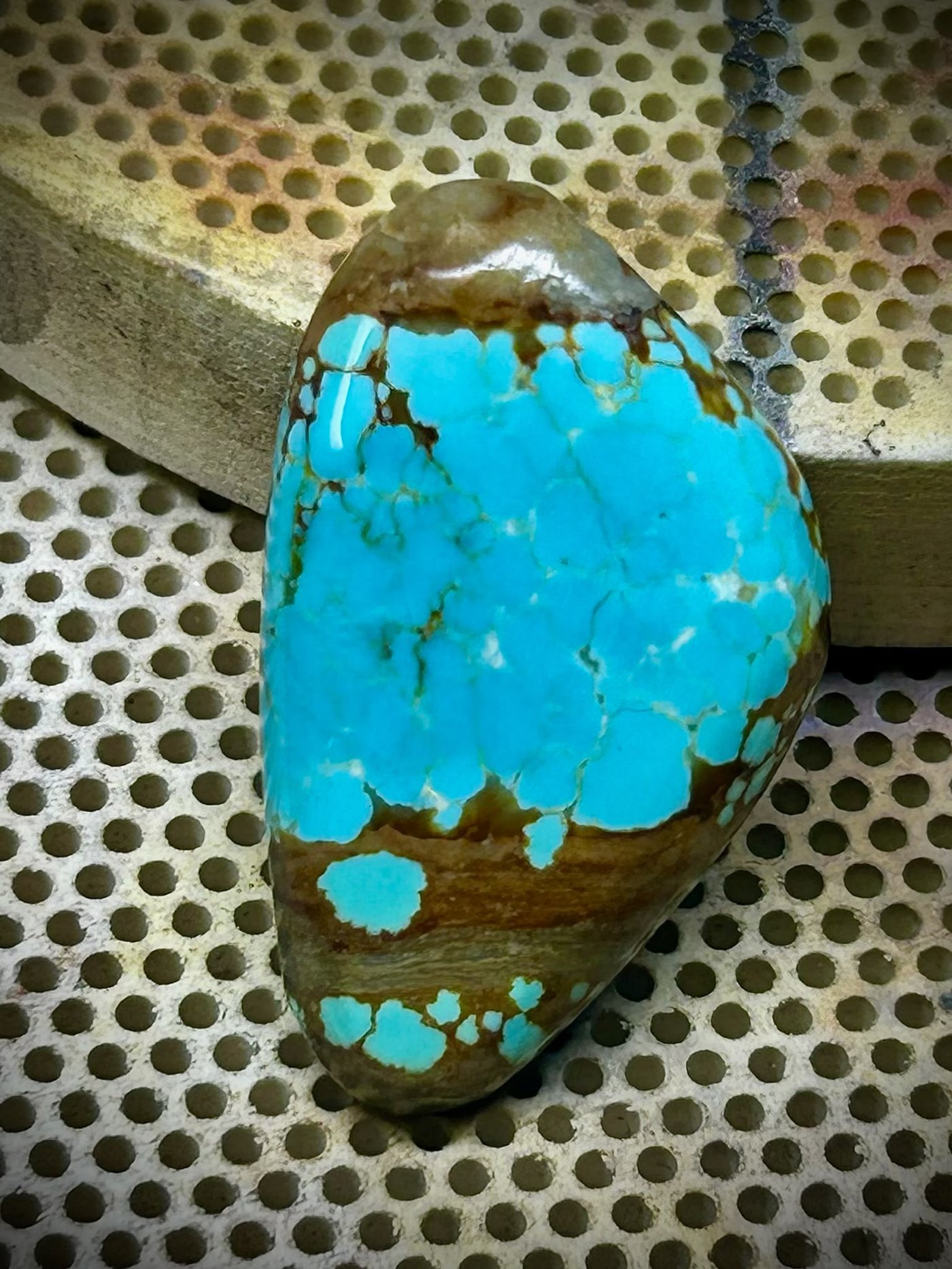 A turquoise cabochon, sky blue with a rust brown web running across it, resting on a perforated bench block.