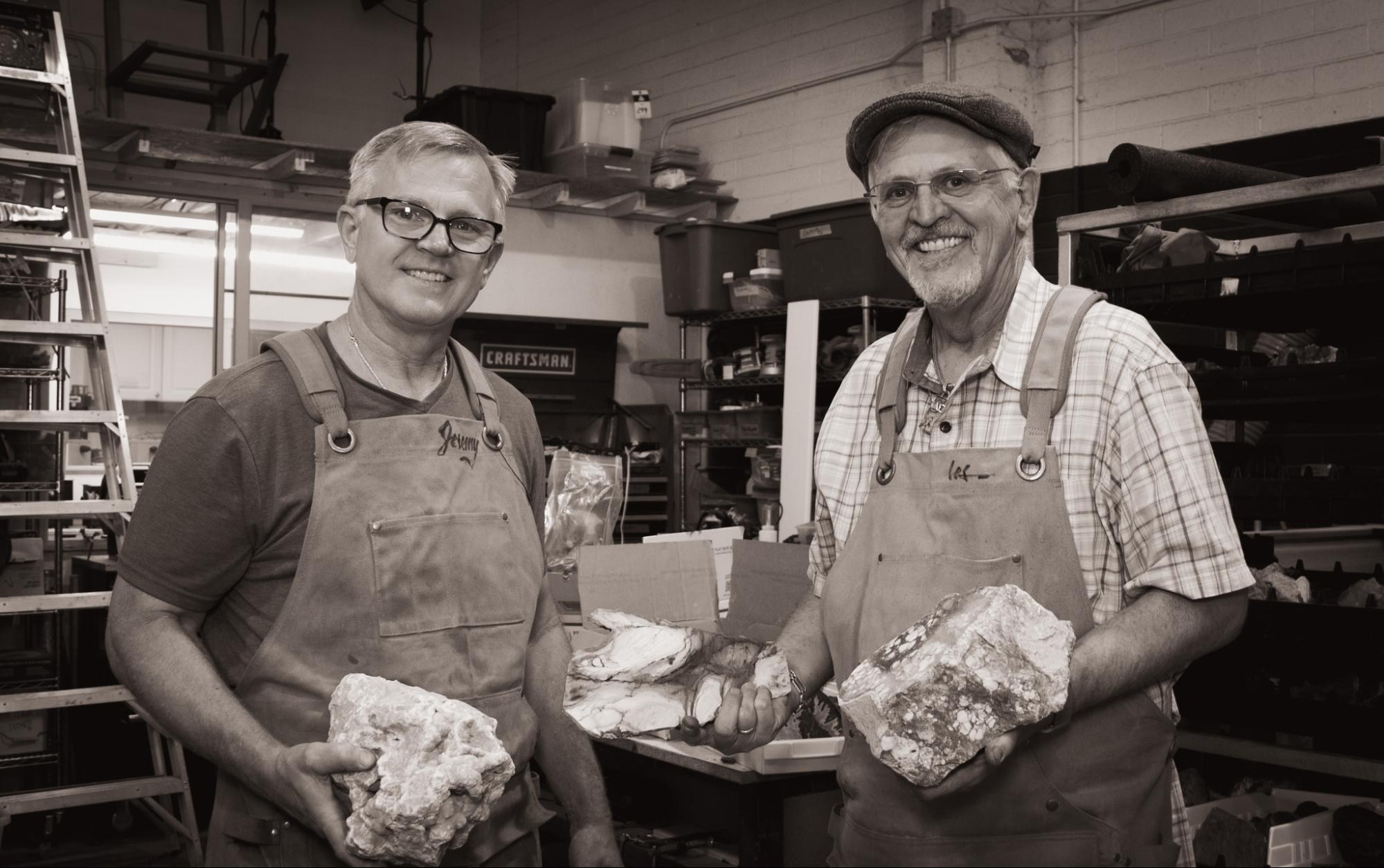 Jeremy and Les in the workshop in their aprons, smiling, each holding a large piece of rough stone.