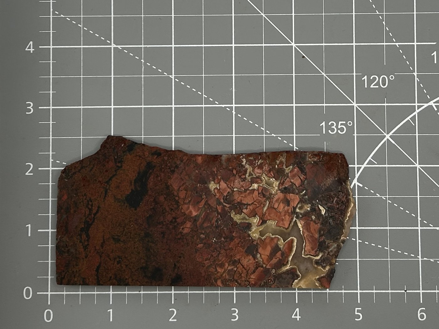 A wild fire jasper slab, deep red and brown with a pale agate edge, lying on the grey cutting mat and measured against its ruler grid.